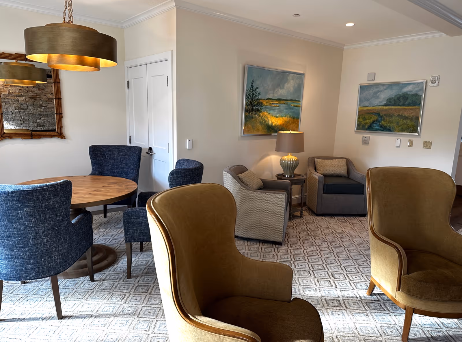A cozy interior room with a round wooden table surrounded by four blue upholstered chairs. Two mustard yellow armchairs are in the foreground. The room has a patterned carpet and cream-colored walls adorned with two landscape paintings. A table lamp sits on a small round side table between two gray armchairs in the corner. A large mirror with a wooden frame hangs on the wall near the table.