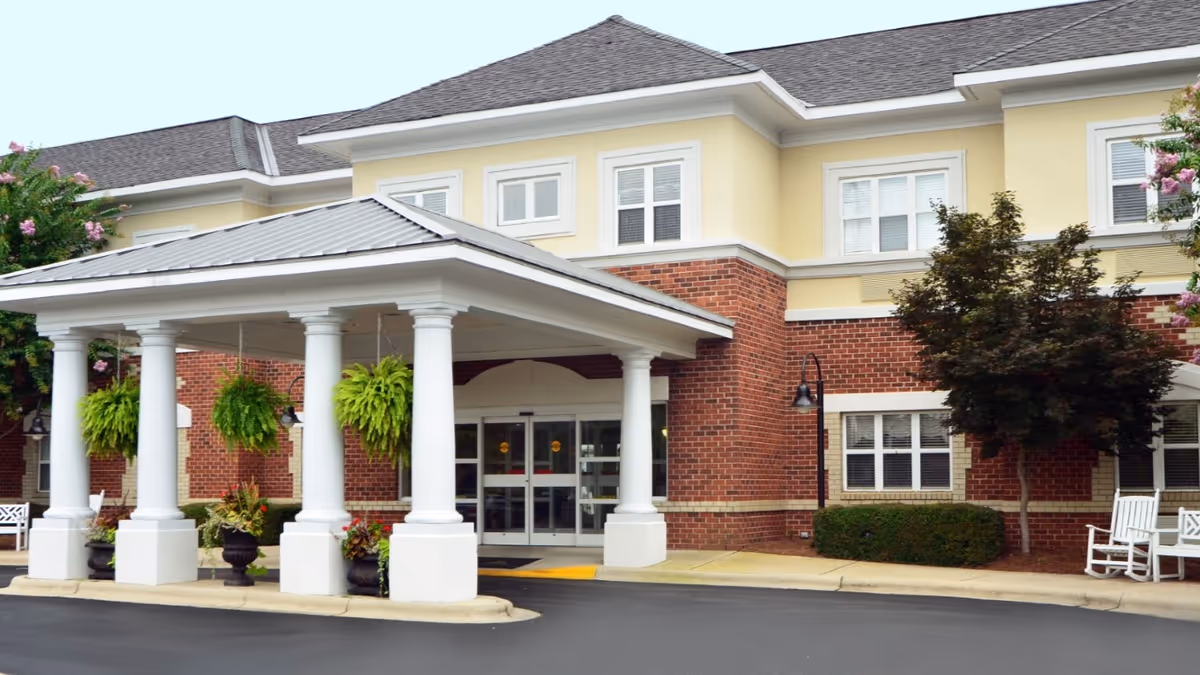 Front exterior view of Morningside of Gastonia building with a covered entrance supported by white columns, hanging green plants, brick and yellow siding, and white rocking chairs near the entrance.