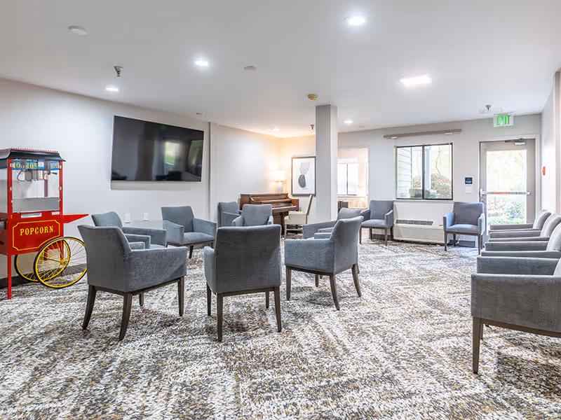 A spacious lounge area with multiple gray upholstered chairs arranged in small groups on a patterned carpet. A red popcorn machine is positioned on the left side near a wall-mounted flat-screen TV. In the background, there is a piano with a lamp and framed artwork on the wall. Large windows and a glass door allow natural light into the room.