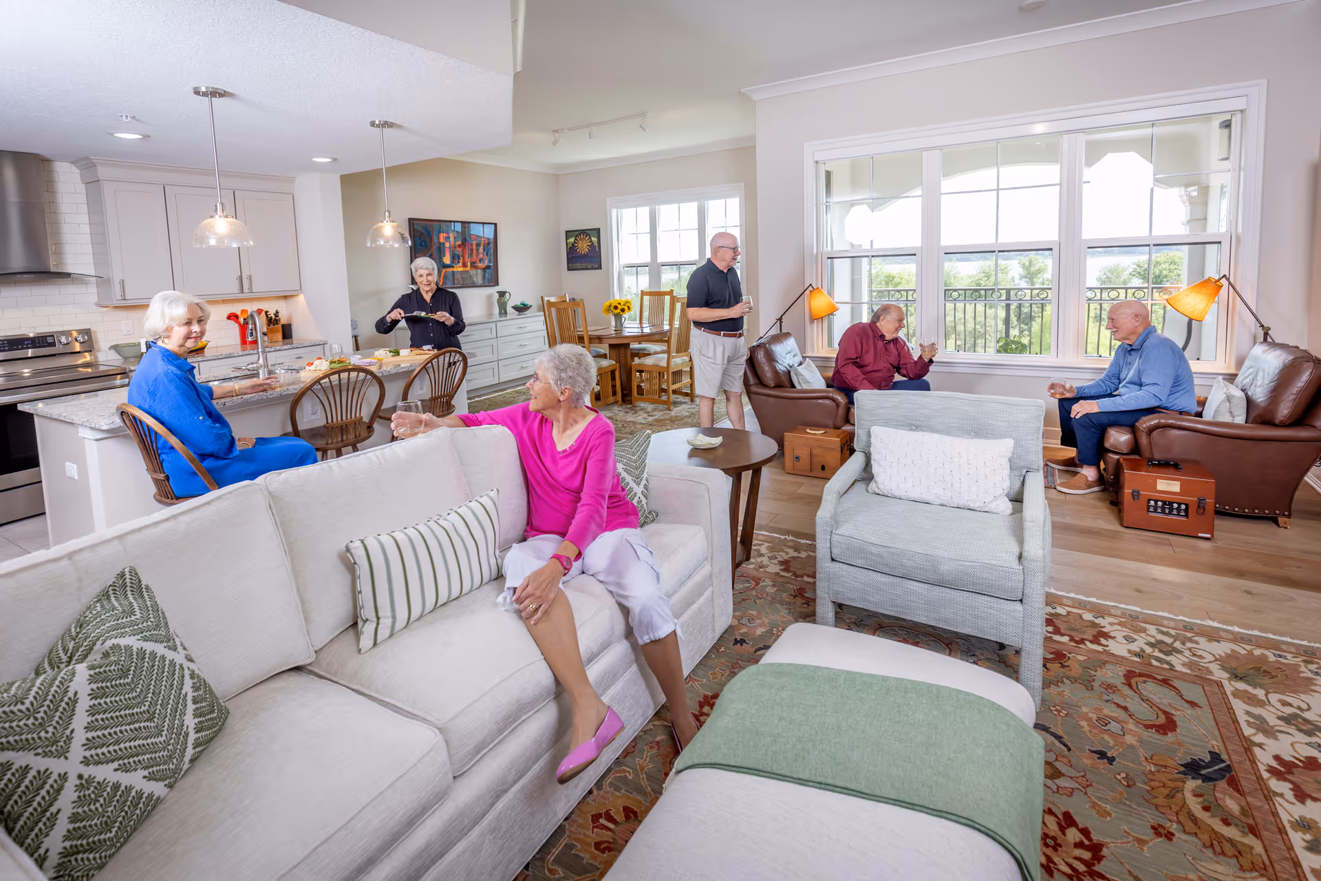 Open-plan living room and kitchen in a senior living facility with several older adults seated and conversing on sofas and at the counter.