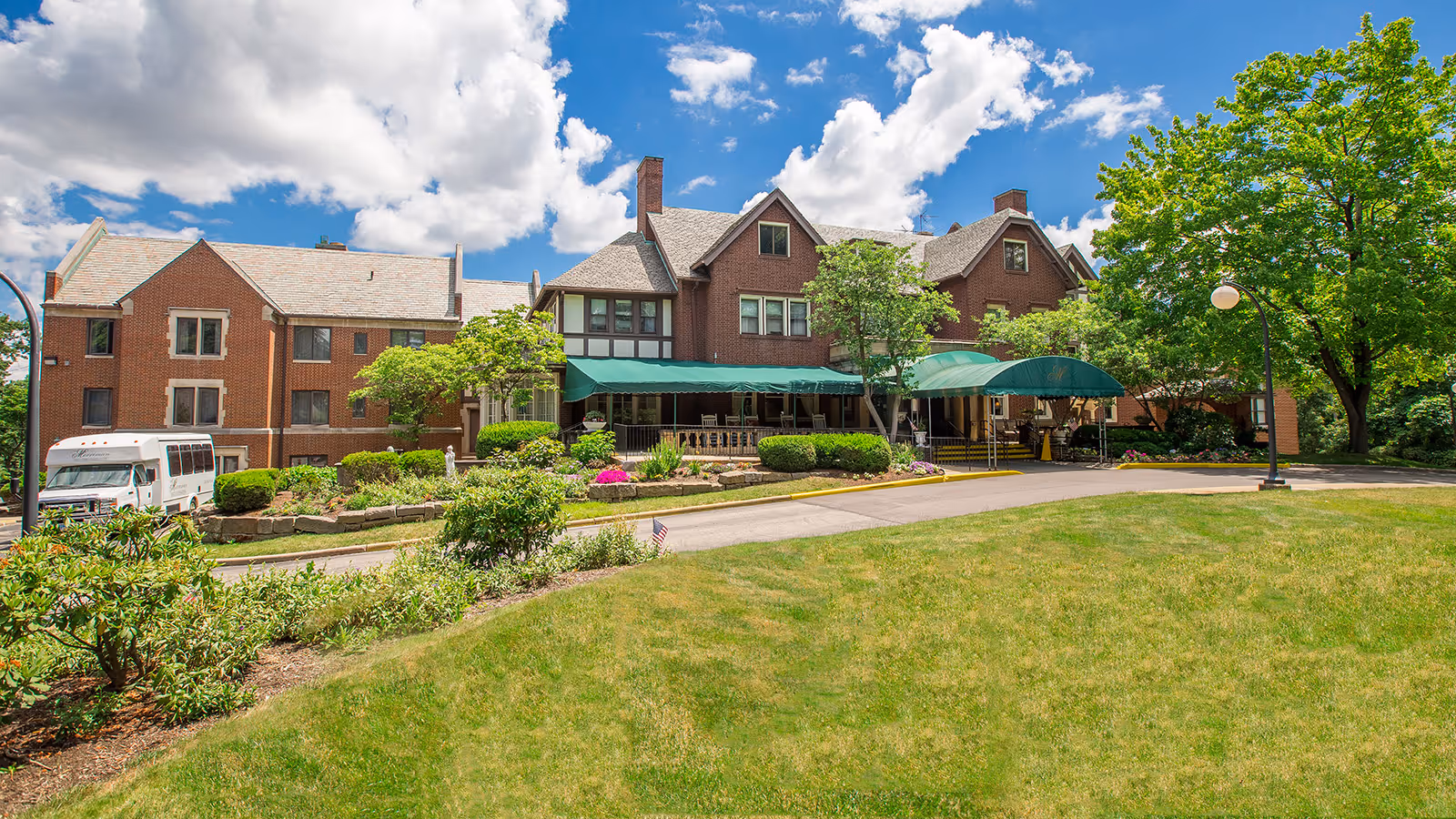 Exterior view of The Merriman facility showing a large brick building with multiple windows and green awnings over the entrance. The building is surrounded by well-maintained landscaping including bushes, trees, and a grassy lawn under a partly cloudy blue sky.