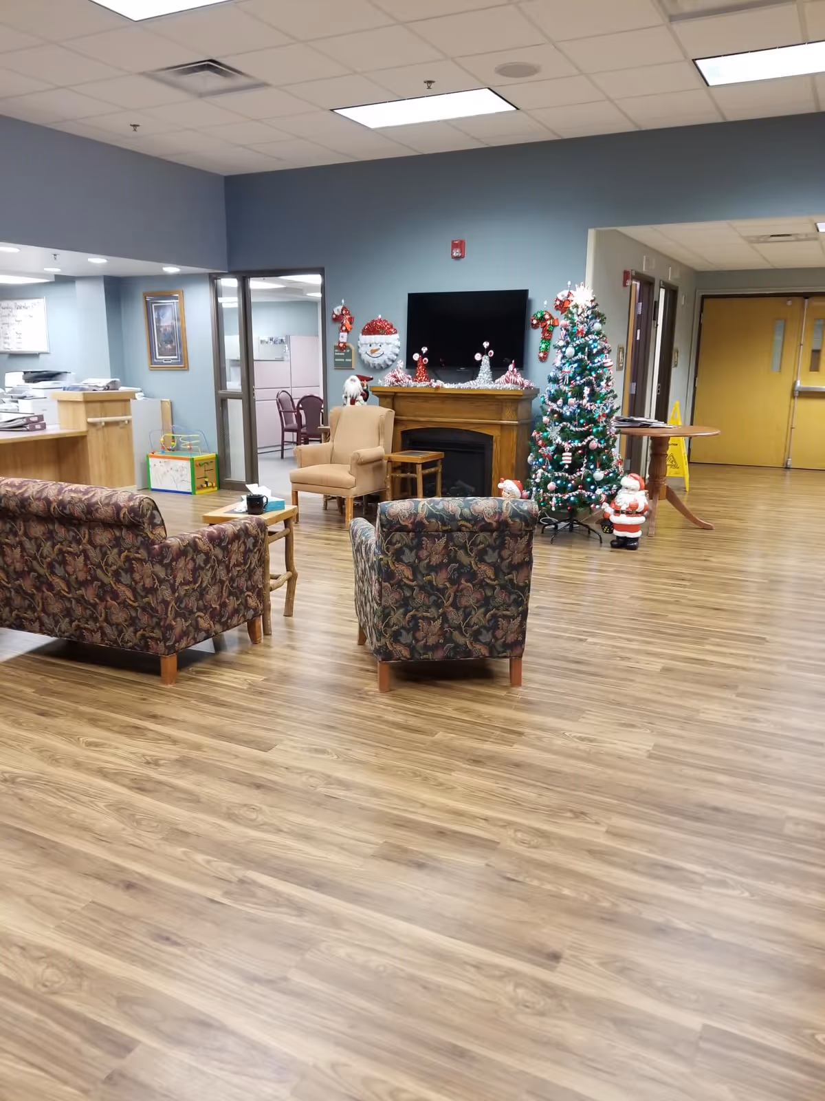 A cozy common area in a senior living facility decorated for Christmas with a Christmas tree, Santa figurines, and festive decorations on a fireplace mantel. The room has patterned armchairs and a sofa, a wooden floor, and a wall-mounted TV. There is a reception desk area to the left and double doors in the background.