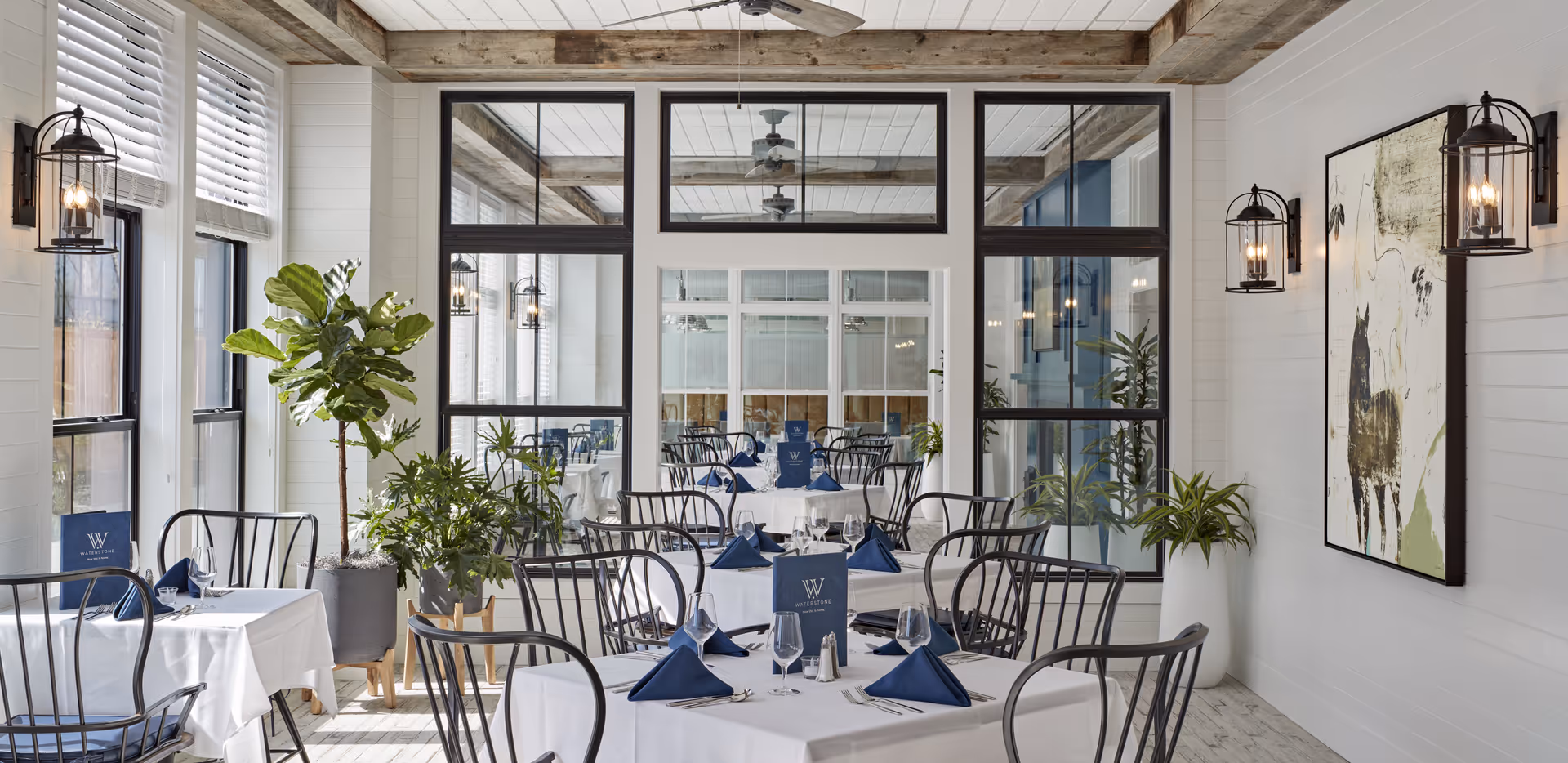 A bright dining room with several tables covered in white tablecloths, each set with blue napkins, glassware, and silverware. The room features large windows with white blinds, black metal chairs, potted plants, wall-mounted lantern-style lights, and a large abstract painting on the right wall. The back wall has large glass panels and doors, reflecting the room and adding depth.
