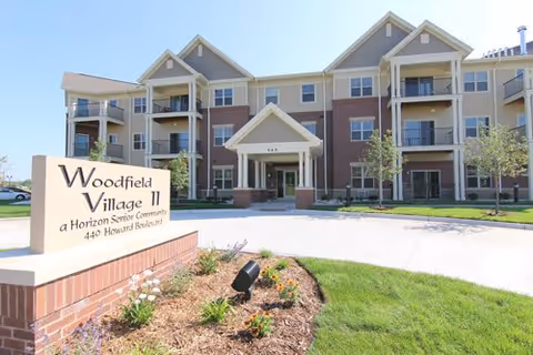 Exterior view of Woodfield Village II, a multi-story senior living community building with balconies, a covered entrance, and landscaped grounds with flowers and grass.