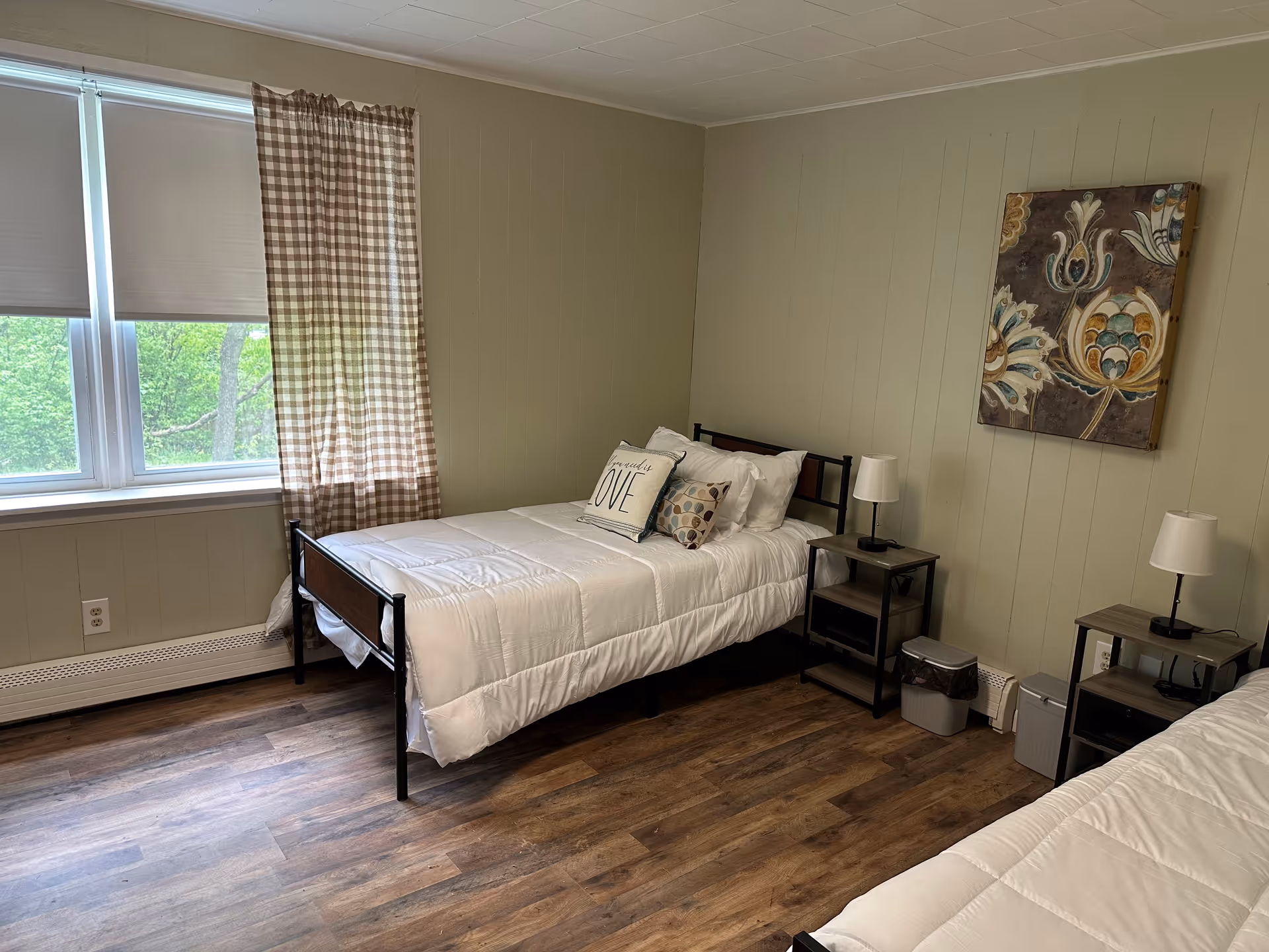 A bedroom with two single beds, each with white bedding and decorative pillows. The room has light green walls, wood flooring, two small bedside tables with lamps, a window with checkered curtains, and a floral painting on the wall.