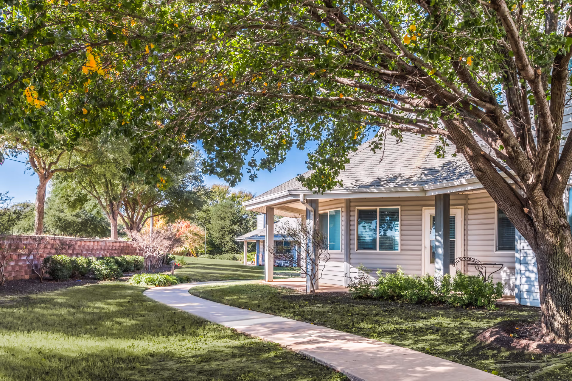 A tree-shaded walkway curves past lawns and landscaping to the front porch of a single-story senior living building.