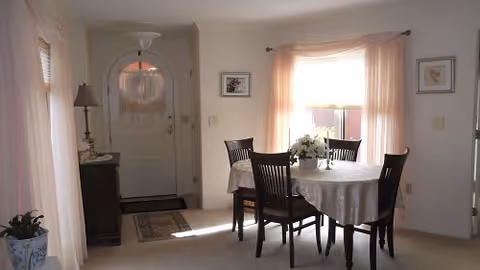 A bright dining area with a round table covered by a white tablecloth and four dark wooden chairs. A floral centerpiece is on the table. The room has light-colored walls, a window with sheer pink curtains, and a white front door with a decorative glass panel. A small cabinet with a lamp and decorative items is near the door.