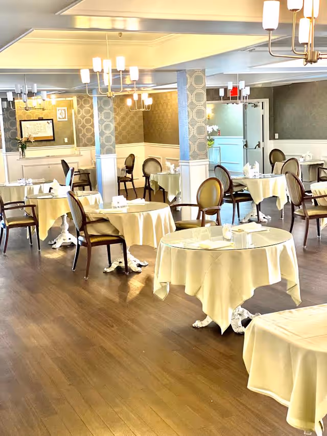 A bright and elegant dining room with round tables covered in white tablecloths, each set with napkins and glassware. The room features wooden chairs with cushioned seats, patterned wallpaper, and modern chandeliers hanging from the ceiling. There are decorative columns and a sideboard with flowers and framed artwork in the background.