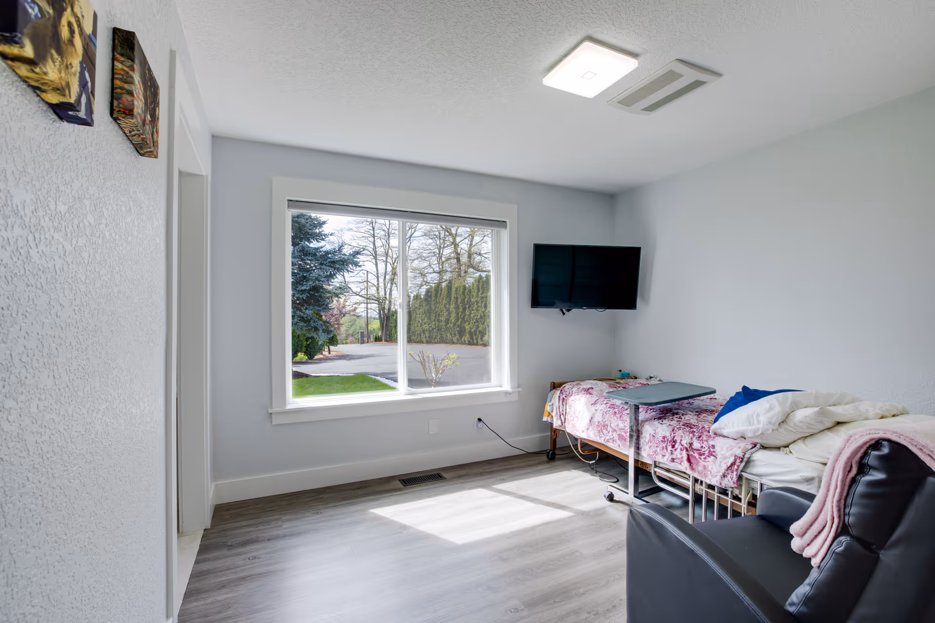 Sunlit single-occupancy bedroom with a window, wall-mounted TV, bed with a floral blanket and an overbed table.