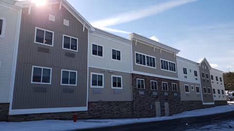Exterior view of a multi-story building with a combination of gray and white siding and stone accents at the base, under a clear blue sky with sunlight shining from the upper left corner.