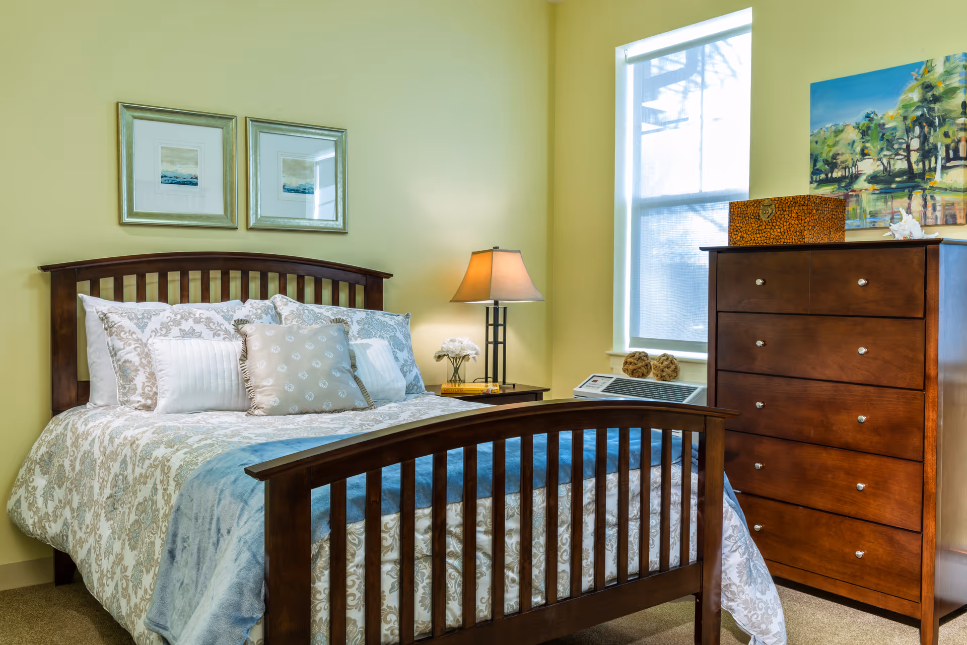 A cozy bedroom with a wooden bed frame featuring a patterned bedspread and multiple pillows. There is a wooden nightstand with a lamp and a small flower arrangement next to the bed. On the right side, there is a wooden dresser with a decorative box and a painting above it. A window with blinds allows natural light into the room.