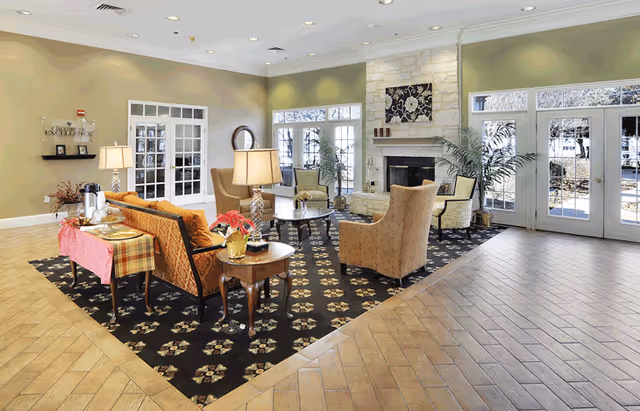 A spacious and well-lit living room area with beige walls and large windows letting in natural light. The room features a patterned dark carpet with floral designs, several upholstered armchairs, a sofa with a plaid throw, wooden side tables with lamps, a stone fireplace, and potted plants near the windows.