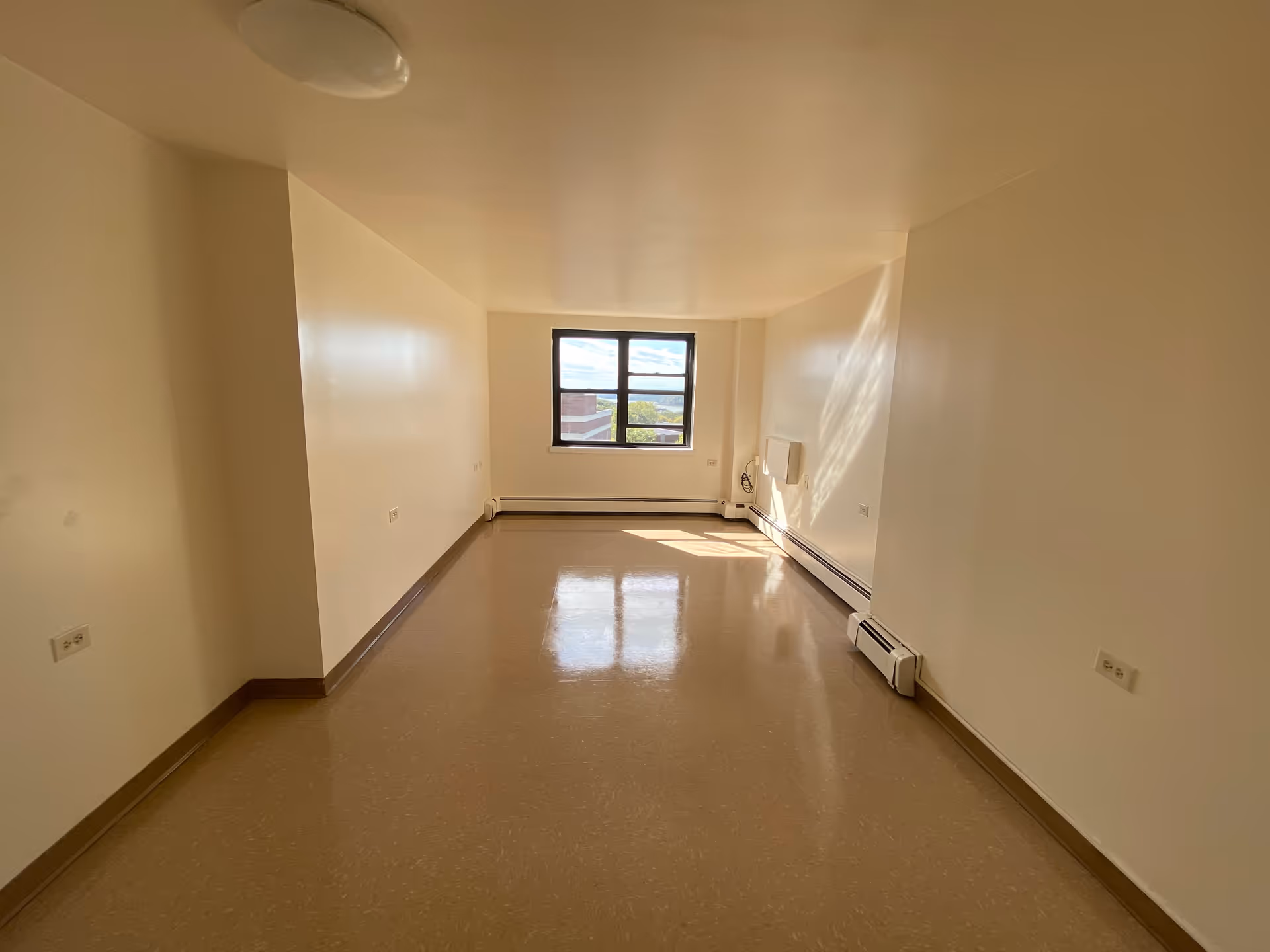 Empty rectangular room with beige walls and floor, a window at the far end letting in natural light, and baseboard heaters along the walls.