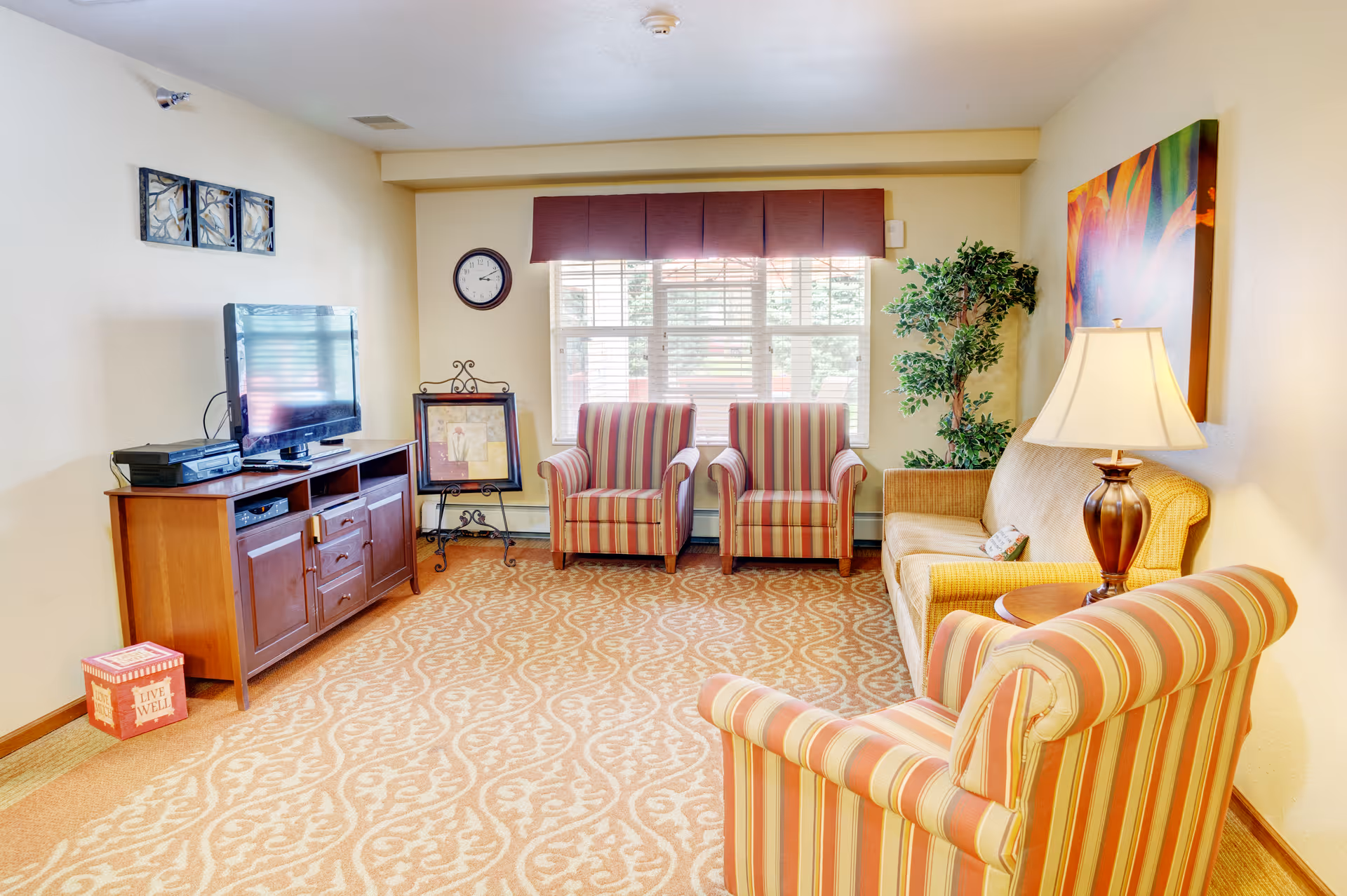 Well-lit living room with striped armchairs, a sofa, TV on a wooden console, lamp, and a window with blinds.