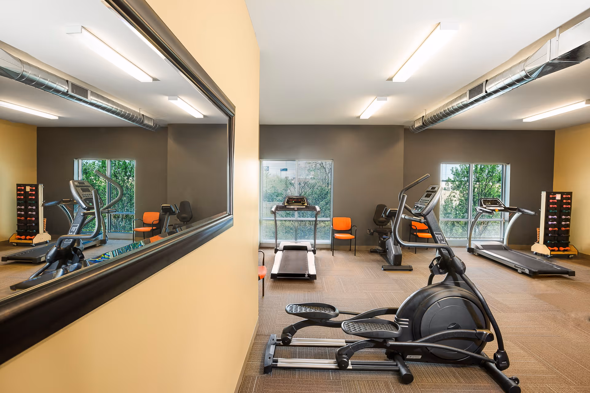 A fitness room with exercise equipment including an elliptical machine, treadmills, stationary bikes, and a rack of dumbbells. The room has large windows letting in natural light, beige and gray walls, and a large mirror on one side reflecting the equipment.
