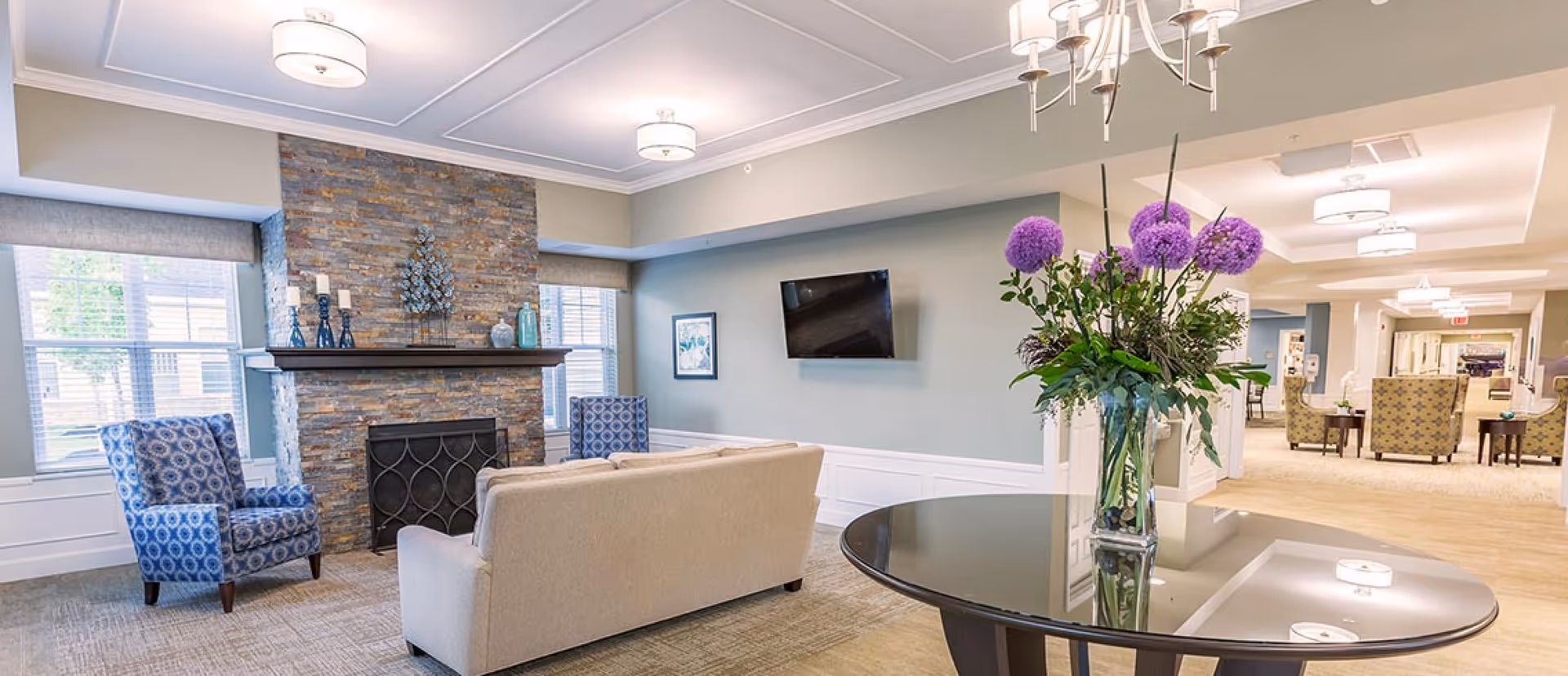 Bright communal living room with seating arranged around a stone fireplace, a wall-mounted TV, and a round table holding a vase of purple flowers.