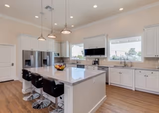 Bright modern kitchen with white cabinetry, a large island with four black bar stools, stainless steel refrigerator, built-in microwave, and two windows above the sink providing natural light.