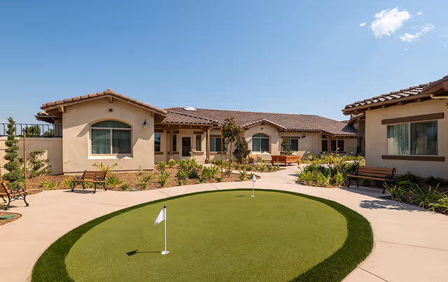 Outdoor courtyard area of a senior living facility with a small putting green in the center, surrounded by a paved walkway, benches, plants, and single-story buildings with tiled roofs under a clear blue sky.