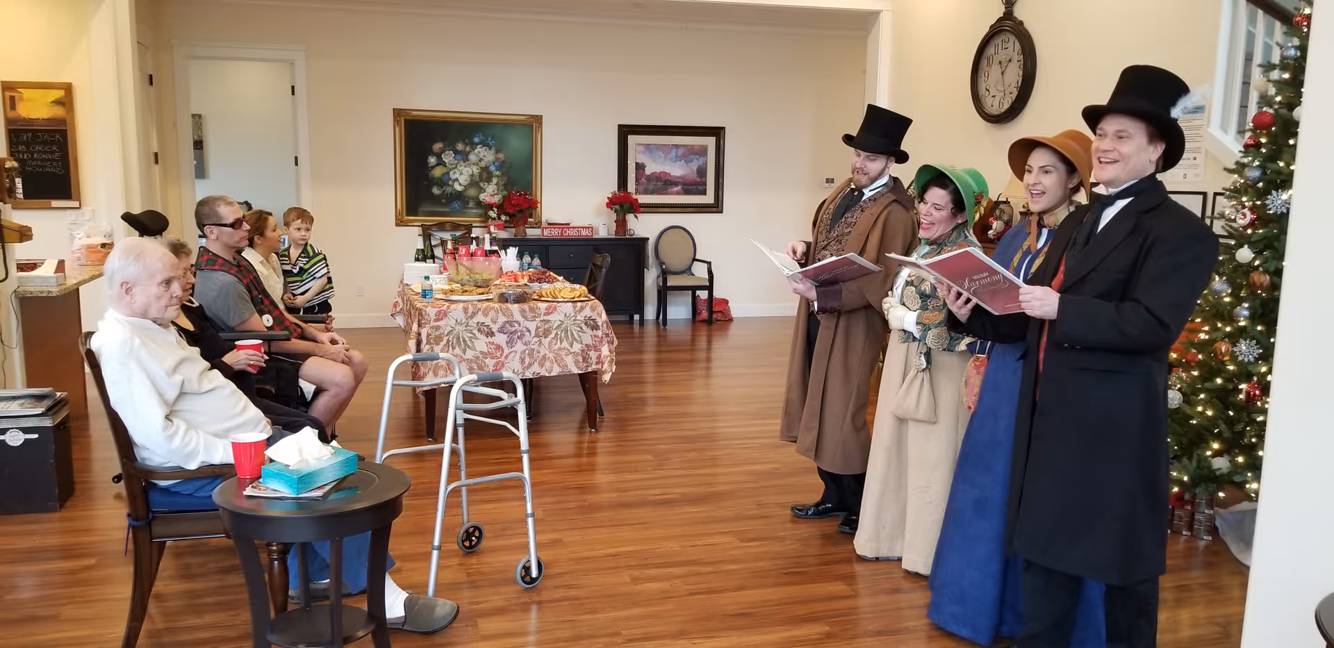 A group of elderly residents and a young boy seated in a common area watching four people dressed in Victorian-era costumes singing Christmas carols near a decorated Christmas tree. A table with snacks and drinks is set up in the background, and the room has wooden flooring and framed paintings on the walls.