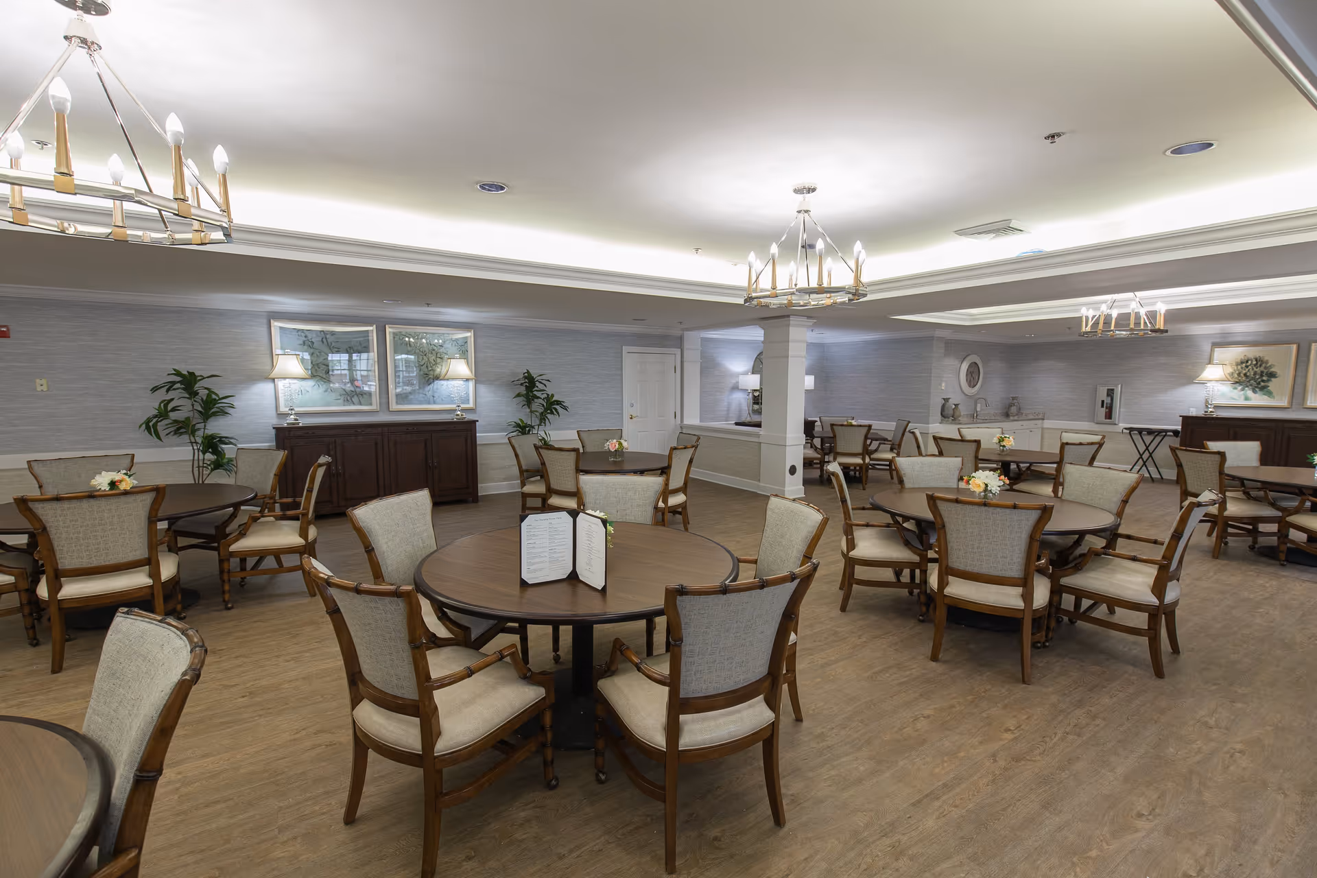 A spacious dining room with multiple round wooden tables surrounded by cushioned chairs. The room features soft lighting from chandeliers and table lamps, light-colored walls with framed artwork, and a wooden floor. Some tables have small floral centerpieces and a menu stand.