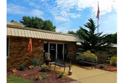 Exterior view of a single-story brick building with a metal roof, a concrete walkway leading to the entrance, landscaped garden beds with shrubs and mulch, two benches near the entrance, and an American flag on a flagpole against a partly cloudy sky.