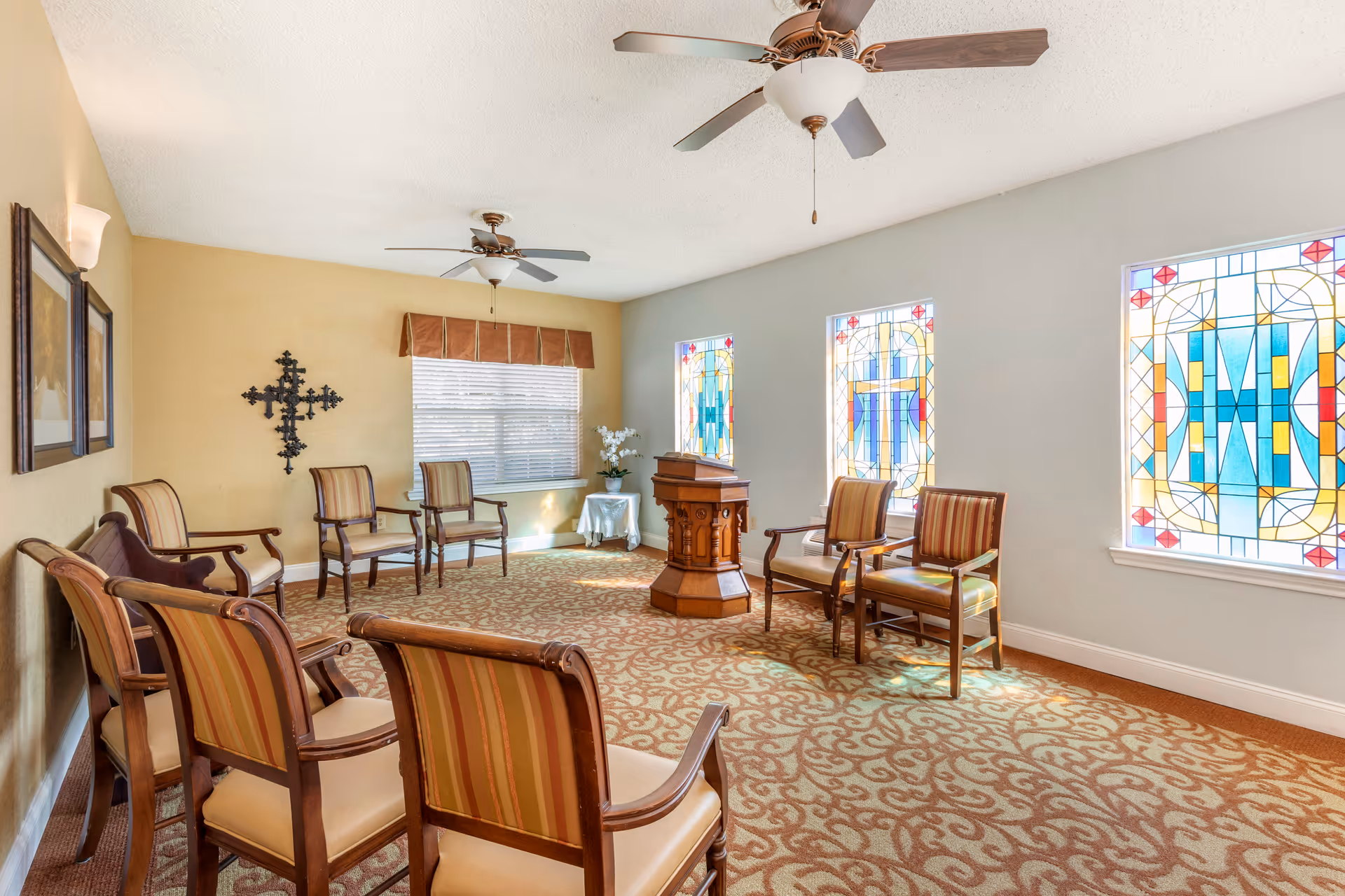 A small chapel or meditation room with several wooden chairs arranged in a semicircle around a wooden podium. The room has patterned carpet, two ceiling fans, stained glass windows with cross designs, a wall-mounted decorative cross, and framed pictures on the wall.