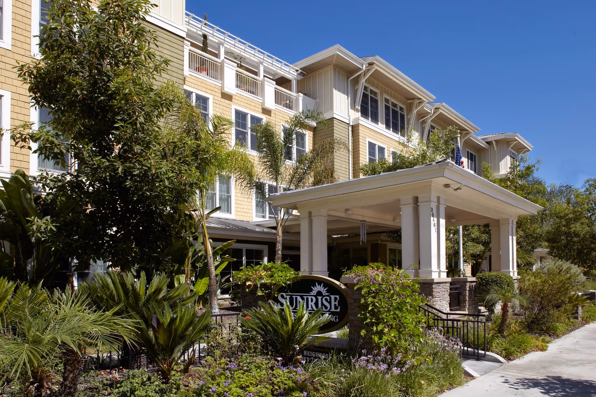 Exterior view of a multi-story senior living facility with beige and white siding, large windows, and a covered entrance supported by columns. The entrance area is surrounded by lush greenery, including palm trees and various shrubs, under a clear blue sky.