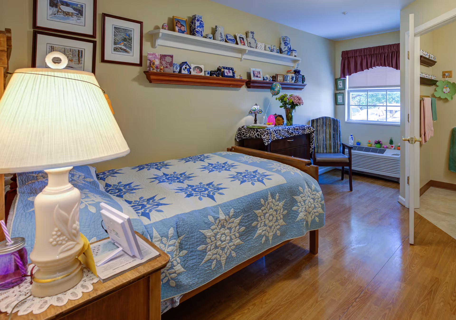 A cozy bedroom with a wooden bed covered by a blue and white quilt featuring floral patterns. A bedside table with a decorative lamp and framed photos is next to the bed. The walls have framed pictures and shelves displaying various decorative items and family photos. A wooden dresser with a floral cloth and a chair are near a window with a maroon valance. The floor is wooden, and an open door leads to a bathroom area with towels hanging.