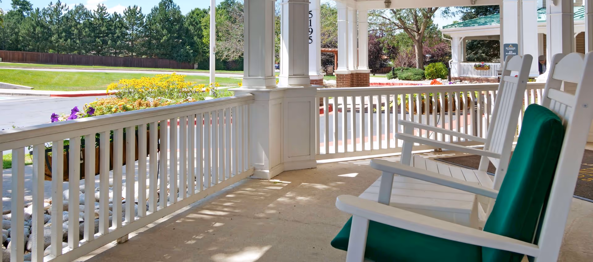 A sunny porch area with white wooden rocking chairs featuring green cushions, white railings, and columns. There are colorful flowers and green trees visible beyond the porch, along with a driveway and a building entrance.