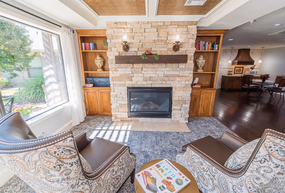 A cozy living room area with two patterned armchairs facing a stone fireplace with a wooden mantel. Built-in wooden bookshelves flank the fireplace, holding books and decorative vases. A large window with white curtains lets in natural light, and a small round table with a book on it is placed between the chairs. In the background, a dining area and kitchen with dark wood flooring are visible.