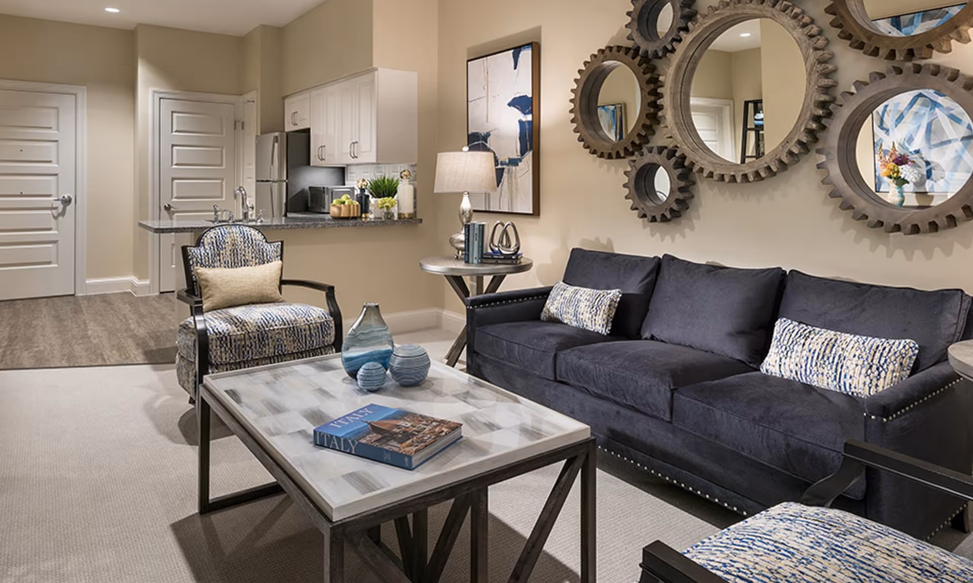A modern living room with a dark blue sofa adorned with patterned cushions, two matching armchairs, a rectangular coffee table with decorative vases and a book titled 'Italy', a side table with a lamp and books, and a wall decorated with multiple round mirrors framed like gears. In the background, a small kitchen area with white cabinets, a refrigerator, and a microwave is visible.