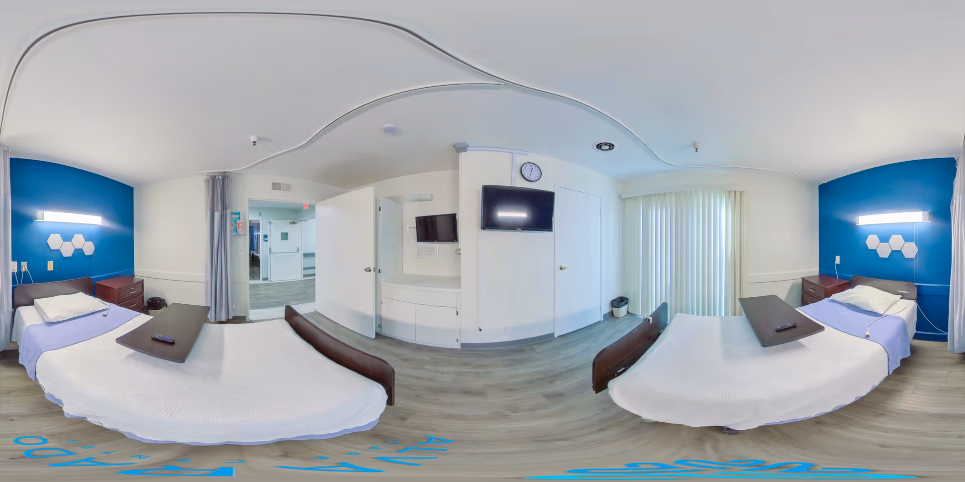 Two single beds in a bright, accessible shared care facility room with blue accent walls, wall-mounted TV, and sliding vertical blinds.