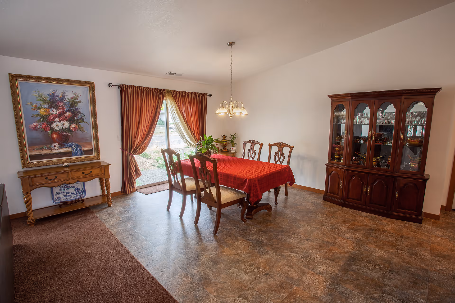 A dining room with a wooden table covered by a red tablecloth and surrounded by four wooden chairs with cushioned seats. There is a wooden cabinet with glass doors displaying glassware on the right side. On the left side, there is a wooden console table with a large framed painting of a floral arrangement above it. A sliding glass door with red and beige curtains lets in natural light.