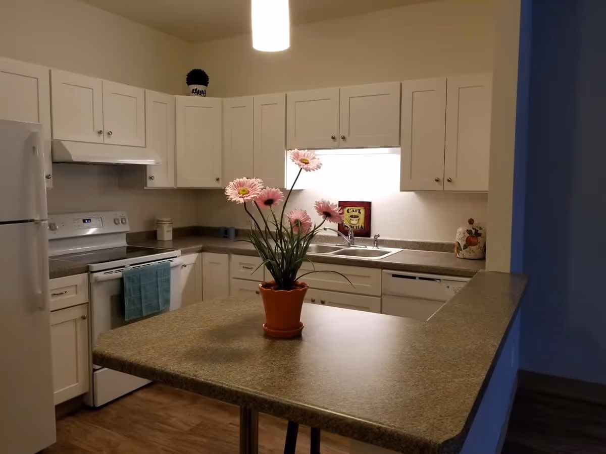 A clean and modern kitchen with white cabinets, a white refrigerator, stove, and dishwasher. There is a countertop with a potted plant featuring pink flowers in the center. The kitchen has a light fixture hanging from the ceiling and a small decorative sign near the sink.