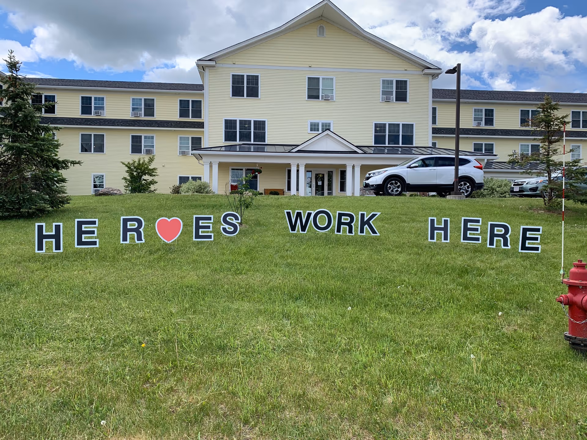 Front of a yellow three-story senior living building with lawn letters spelling 'HEROES WORK HERE' including a red heart and cars parked by the entrance.