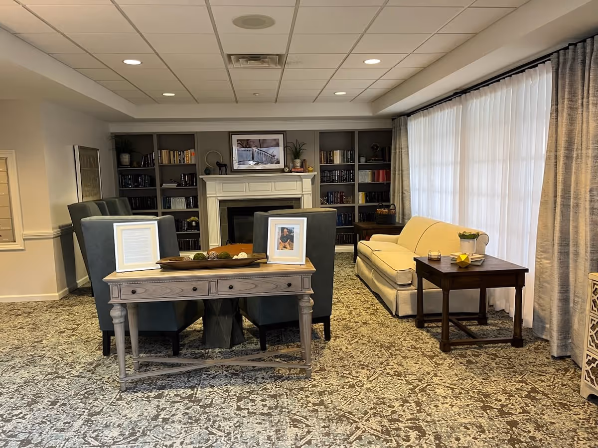 A cozy living room area with a beige sofa, a wooden side table with a plant and decorative items, a wooden console table with framed photos and decorative objects, four upholstered chairs, a fireplace with a mantel, and built-in bookshelves filled with books. Large windows with sheer white curtains and heavier drapes allow natural light into the room.