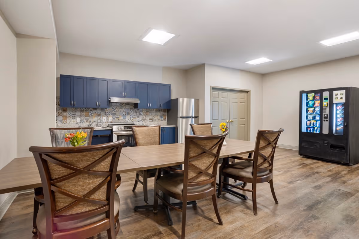 A communal dining area with a long wooden table surrounded by six wooden chairs with cushioned seats. In the background, there is a kitchen area with blue cabinets, a stainless steel refrigerator, stove, and a decorative tile backsplash. To the right, there is a vending machine stocked with snacks and drinks. The room has wood flooring and bright overhead lighting.