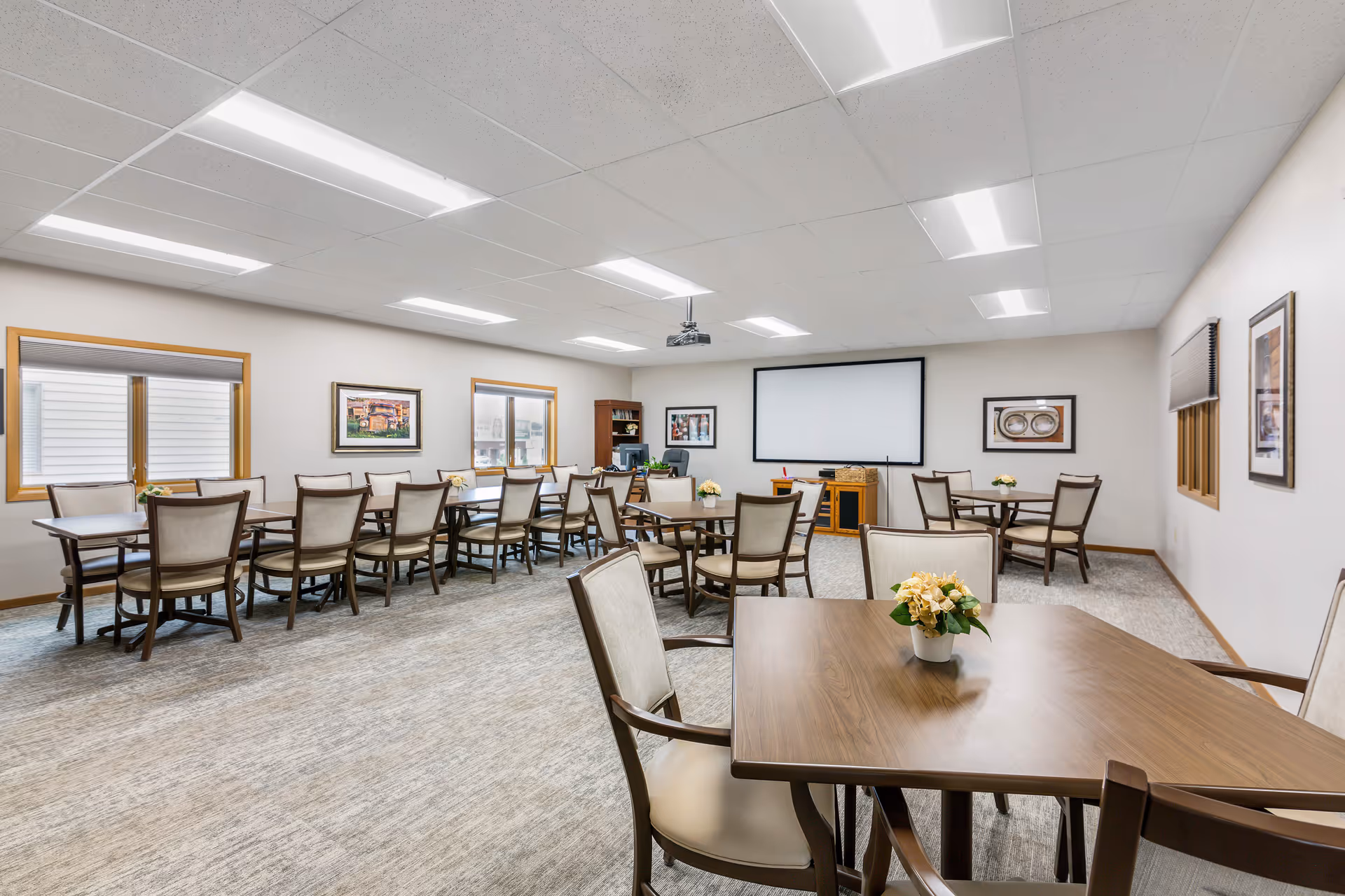A well-lit senior living facility room with multiple wooden tables and cushioned chairs arranged neatly. The room has a projector mounted on the ceiling and a large projection screen on one wall. There are framed pictures on the walls and windows with blinds, and small flower arrangements on the tables.