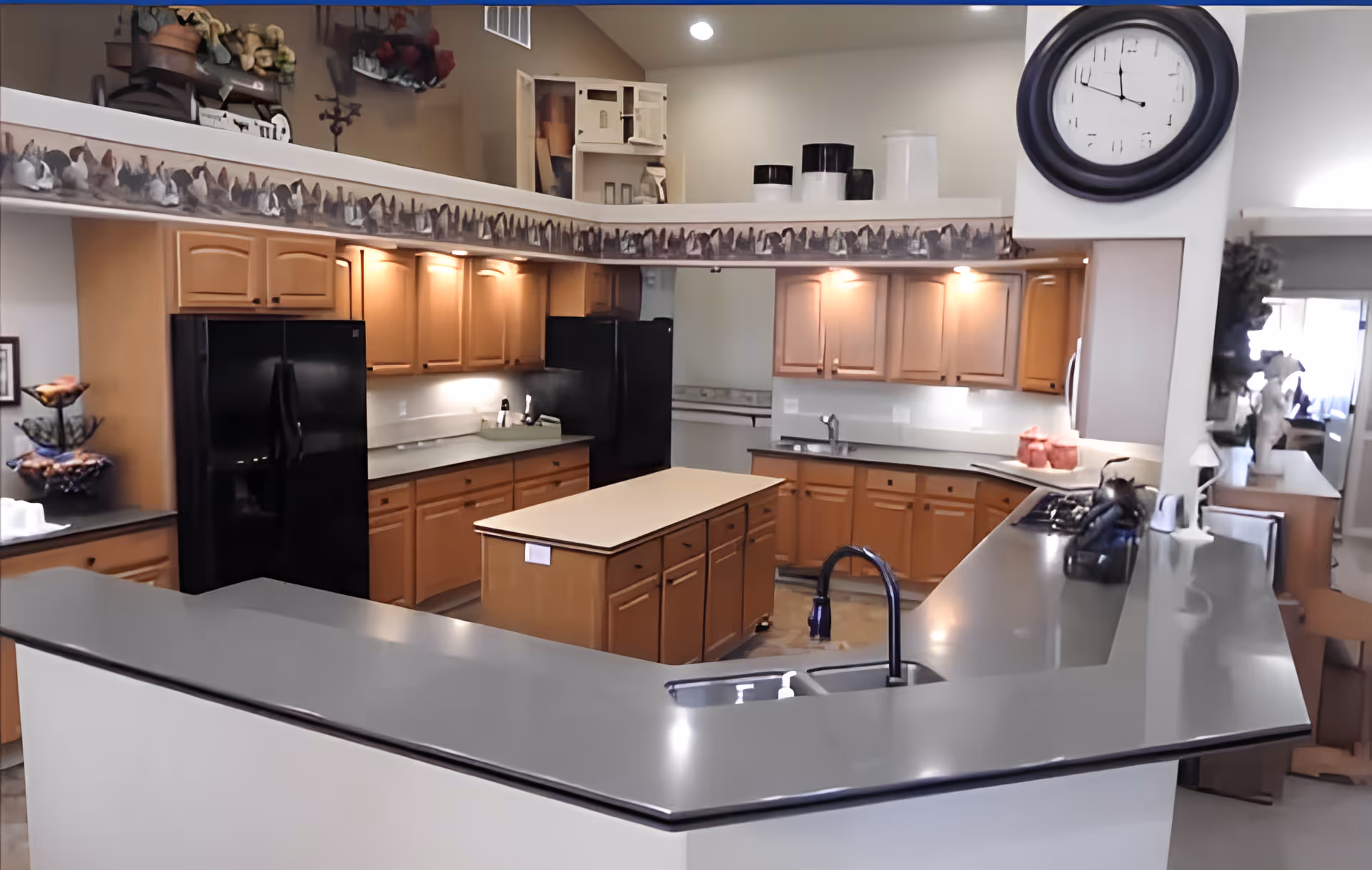 A spacious kitchen with wooden cabinets, black appliances including two refrigerators, a central island, and a large counter with a sink and faucet. The kitchen is well-lit with under-cabinet lighting and ceiling lights. A large black-framed clock is mounted on the wall above the counter. Decorative items and containers are placed on the countertops and shelves.