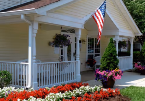 Front porch of a single-story building with white railings and columns, an American flag hanging from the roof, hanging flower baskets, potted plants, and a flower bed with red, white, and purple flowers in front.