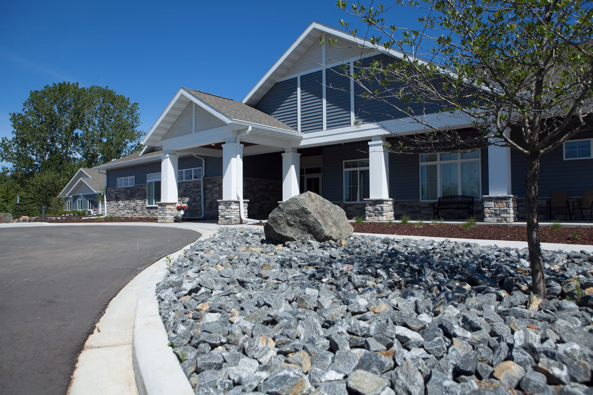 Front exterior of an assisted living building with a covered entrance, stone rock bed, and a curved driveway.