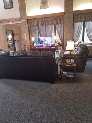 A cozy living room area with a dark leather sofa and recliner facing a television. The TV is on a wooden stand with decorative items around it. The room has large windows with curtains and valances, a side table with a lamp and a telephone, and carpeted flooring. The walls are light-colored with brick columns and framed artwork.