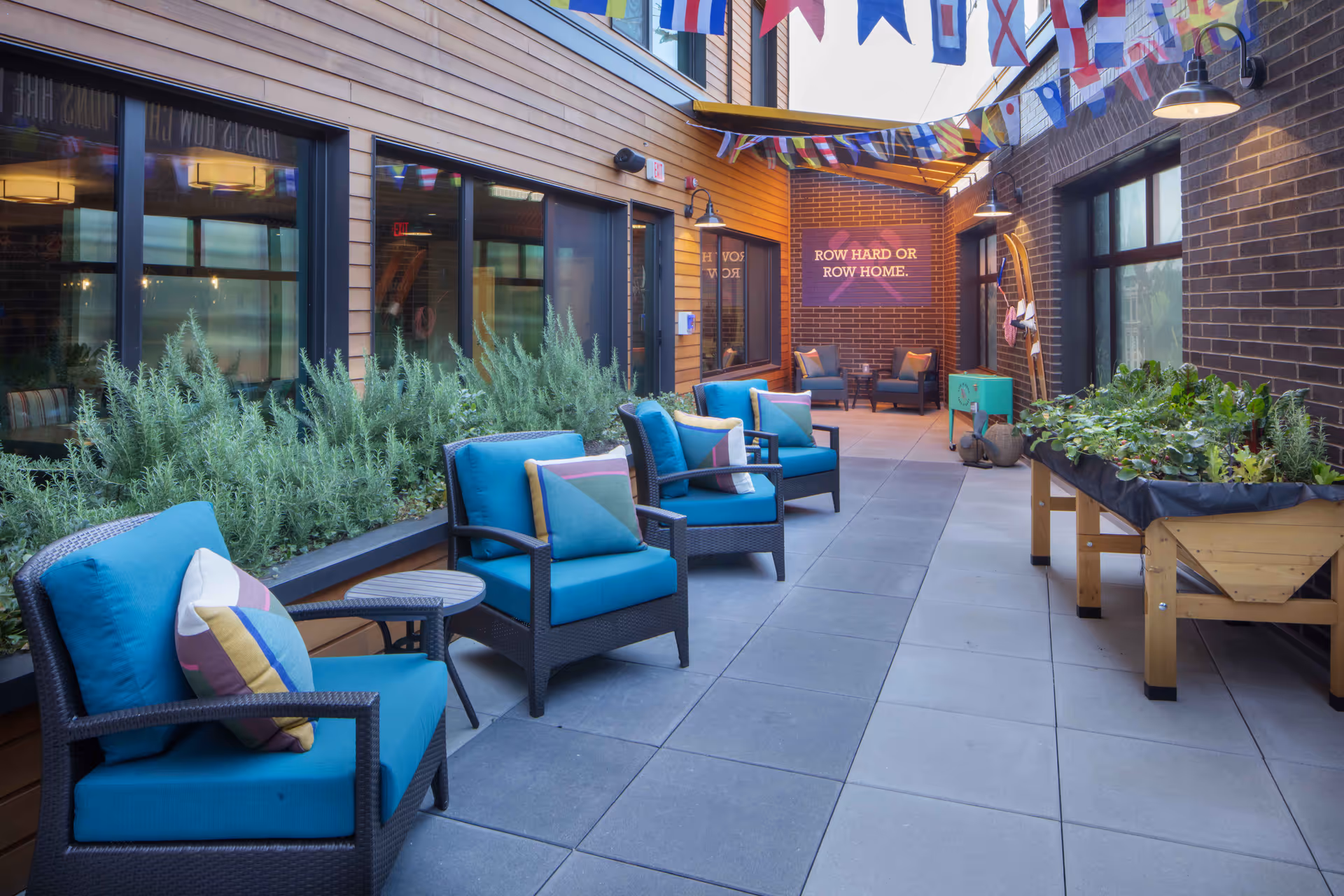 A bright outdoor courtyard with teal cushioned chairs, planters, decorative flags, and a brick wall sign that reads "ROW HARD OR ROW HOME."