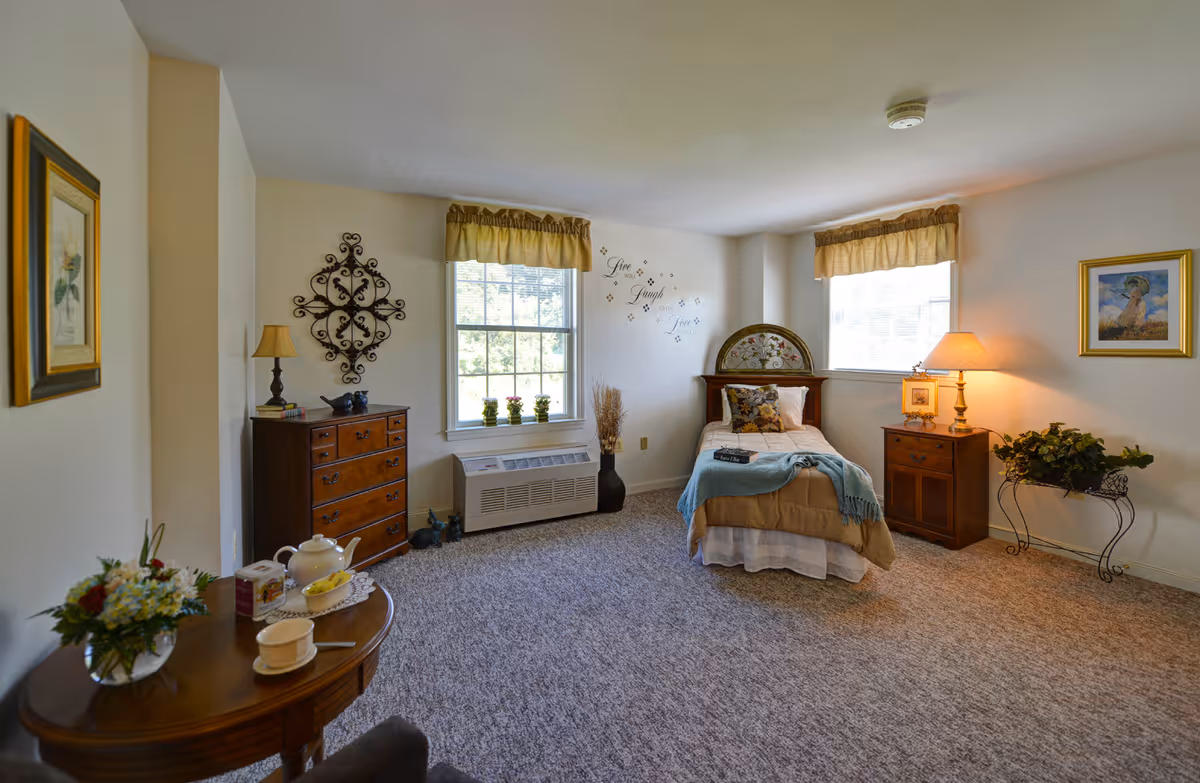 A cozy senior living bedroom with a single bed adorned with pillows and a throw blanket. The room features two windows with yellow valances, a wooden dresser with a lamp and decorative items, a small wooden nightstand with a lamp, framed artwork on the walls, a small table with a tea set and flowers, and a carpeted floor. The wall above the bed has decorative text that reads 'Live Laugh Love.'