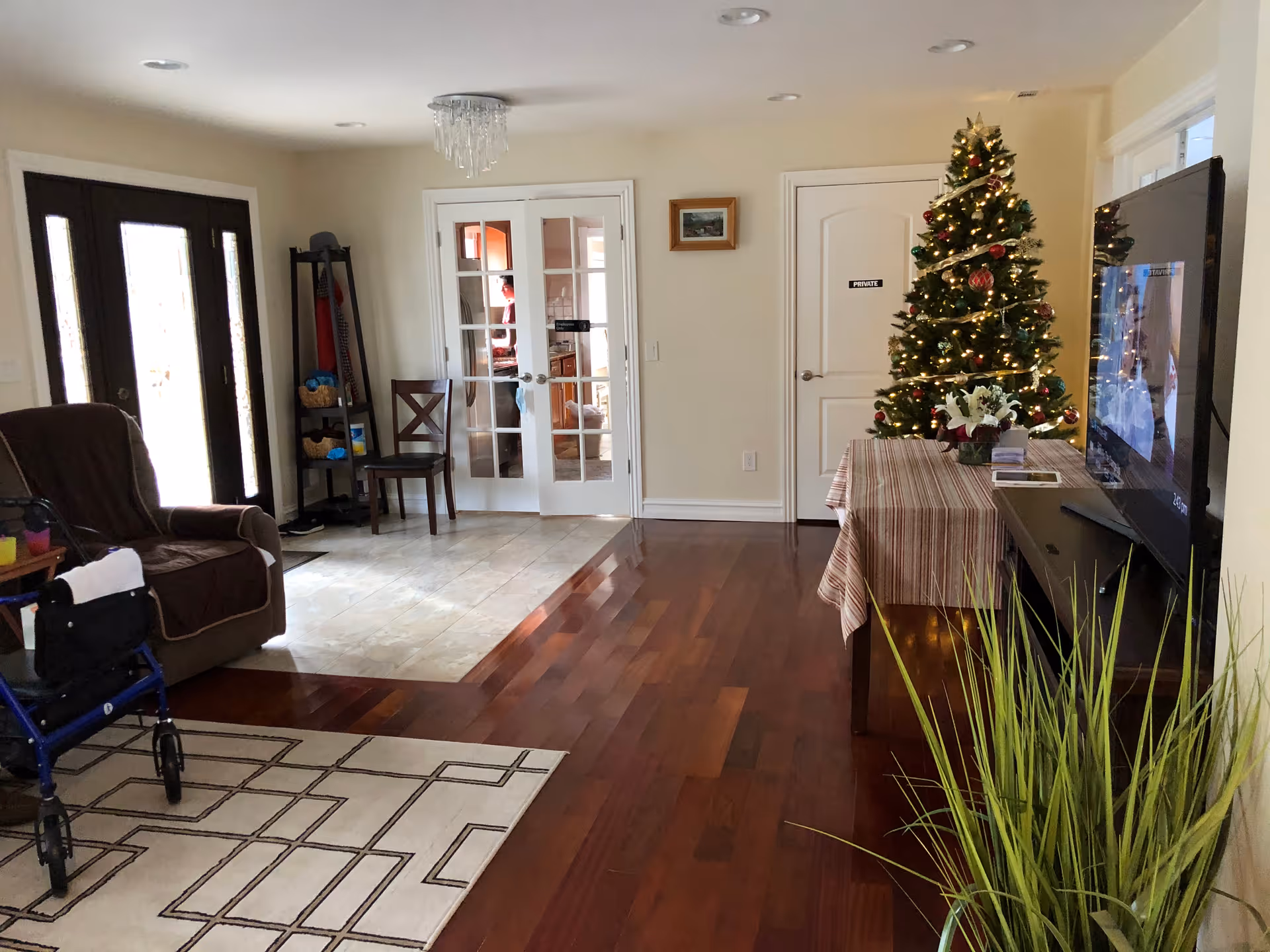 A cozy living room area with wooden flooring and a beige rug with geometric patterns. There is a brown armchair with a walker beside it on the left. Near the back, there are double glass doors leading to another room, and a single door labeled 'PRIVATE'. A decorated Christmas tree with lights and ornaments stands next to a table covered with a striped tablecloth. A flat-screen TV is mounted on the right wall, and a tall green plant is in the foreground on the right side.