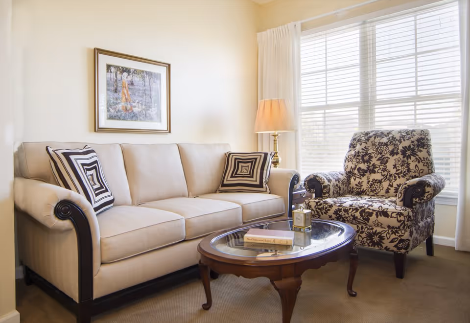 Sunlit living room with a beige sofa, patterned armchair, glass-top coffee table, and a lamp beside a large window.