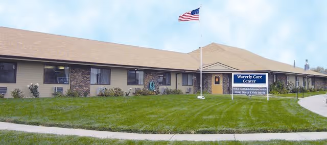 Single-story care facility front with a flagpole, lawn, and a sign reading 'Waverly Care Center'.