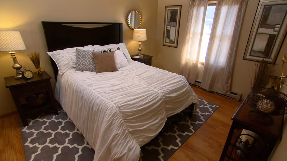 A cozy bedroom with a neatly made bed featuring white ruched bedding and three decorative pillows. The room has wooden floors with a patterned gray and white rug under the bed. There are two dark wooden nightstands on either side of the bed, each with a lamp. A round mirror and framed artwork hang on the walls, and a window with sheer curtains allows natural light to enter the room.