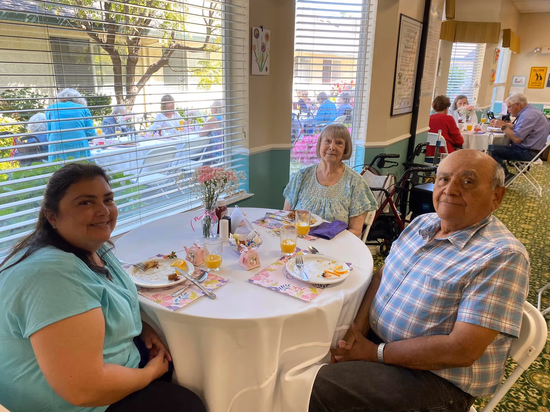 Three adults seated at a round, decorated table enjoying a meal in a bright dining room with large windows showing an outdoor seating area.
