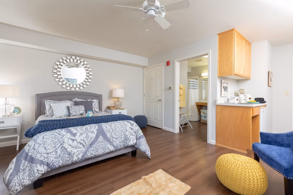 A cozy bedroom in a senior living facility featuring a bed with patterned bedding and multiple pillows, two bedside tables with lamps, a decorative round mirror above the bed, a ceiling fan, and wood flooring. To the right, there is a small kitchenette area with wooden cabinets and a blue armchair with a yellow knitted pouf. An open door reveals a bathroom with a shower curtain and towels.